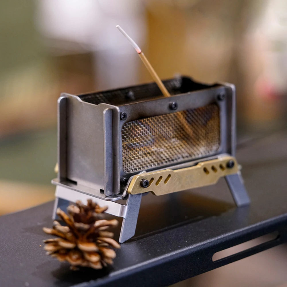 A portable mini stainless steel camping incense burner sitting on a wooden table. The miniature stove design adds a special touch of style and charm, ideal for both home and outdoor use. The product, with its included wood clip and storage bag, is perfect for creating a cozy atmosphere anywhere. Built from high-quality materials, it promises durability and a long lifespan. Please note, slight color and size variations may be present due to lighting effects and manual measurements.